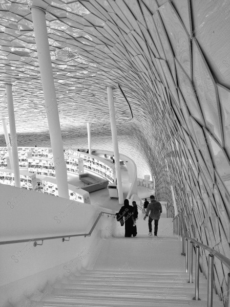 An image of a modern library featuring an innovative architectural design with a geometric patterned ceiling composed of multi-faceted shapes. Several people are walking down a wide white staircase leading to the reading area. The elegant ceiling and white pillars impart a futuristic feel. A couple is visible, one of whom is wearing a black abaya.