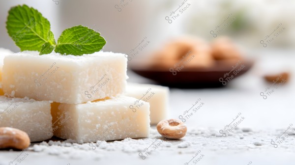 A group of white sugar-dusted cubes decorated with a fresh green mint leaf. Behind them, some circular cookies appear in a blurred wooden plate. The tabletop is sprinkled with some crumbs.