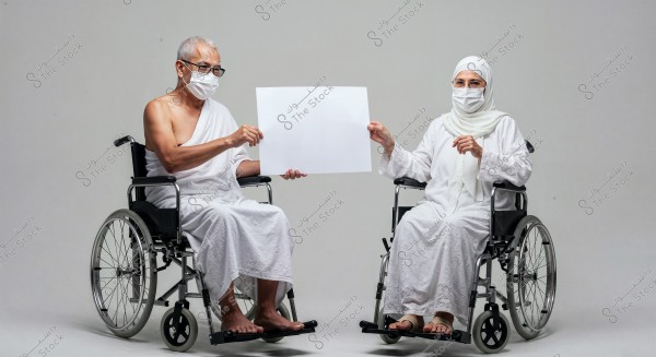 A man and a woman sitting in wheelchairs in a studio, wearing white Ihram garments for performing Umrah or Hajj. The man is bare-shouldered while the woman is wearing a headscarf covering the head and chest. Both are wearing medical masks and are holding a white board between them. The background is plain and white.