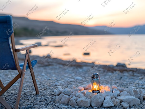 A wooden chair with a blue seat positioned on a pebbled beach. In front of the chair, there is a charming lantern surrounded by large white stones, lit with a flame. In the background, a sunset over the water creates beautiful color gradients in the sky.