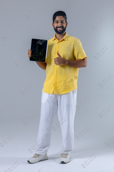 A portrait of a man standing in a studio, wearing a yellow shirt, white pants, and white sneakers. He is holding a tablet in his left hand and giving a thumbs-up with his right hand, smiling. The background is a simple gray color.