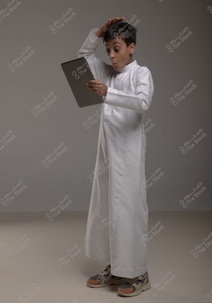 An image of a boy wearing a traditional long white thobe, standing with a surprised expression while holding a tablet in one hand and tilting his head upward with his other hand. The boy appears amazed or astonished. The background is a simple gray and he is wearing athletic shoes.