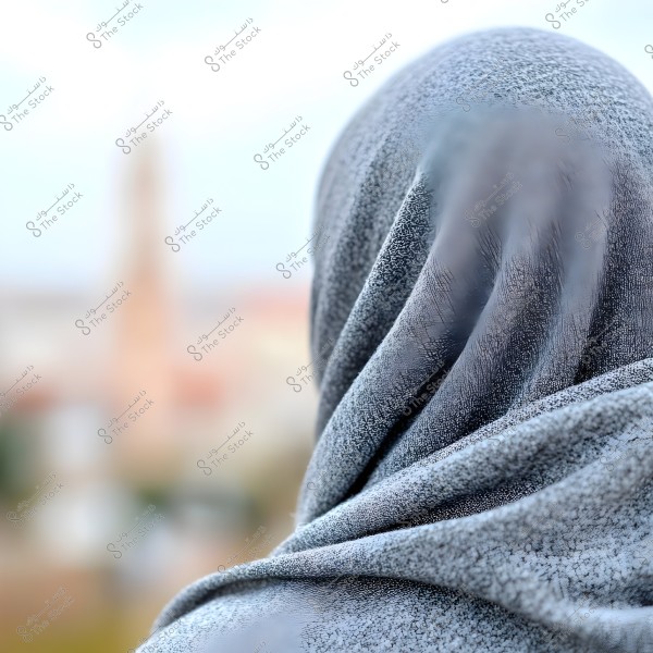 An image showing a person wearing a grey hijab, photographed from the back with a blurred background displaying faded colors and indistinct structures. The focus is on the texture of the hijab.