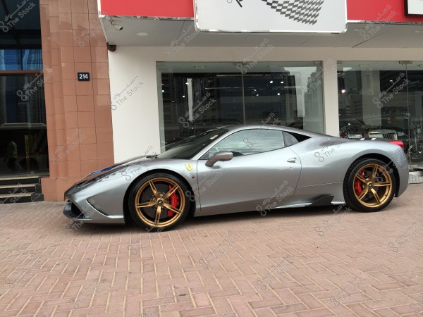 Image of a luxury silver sports car with gold wheels on a brick pavement. The car is parked in front of a modern building with a glass facade, with city landmarks visible in the background.