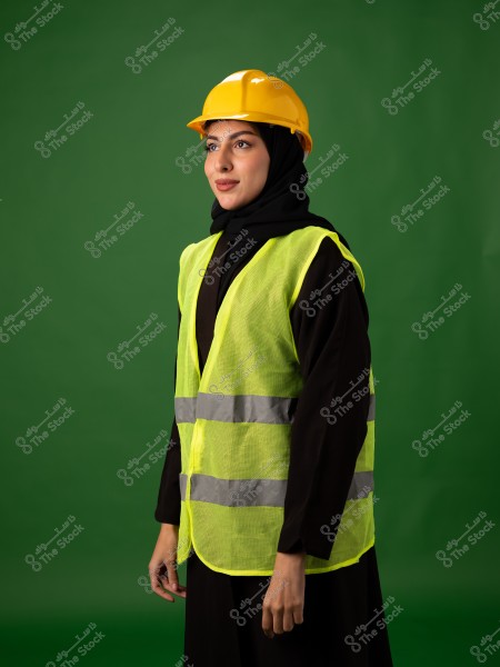 A woman wearing a yellow reflective vest and a yellow construction helmet stands in front of a green background. She is dressed in a black abaya and hijab. She appears in a formal and serious pose, suggesting a professional role in engineering or construction.