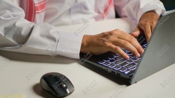 The image shows the hands of a person wearing a white thobe with red trim, working on a laptop with a backlit keyboard. A mouse is next to the laptop on a white table.