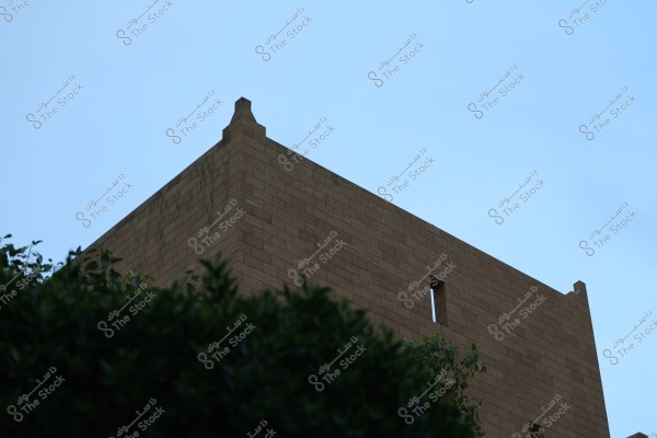 The image shows a portion of a stone building with a brown facade and traditional architectural features, rising towards the blue sky. Some green trees are visible at the bottom.