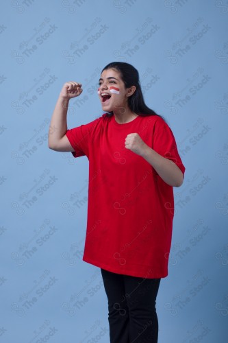 Portrait of a Saudi woman wearing a red T-shirt cheering the football team on a blue background and showing expressions of joy and enthusiasm, World Cup.