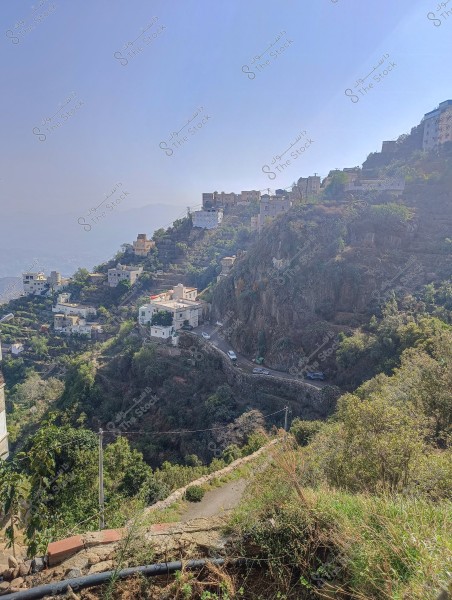 An image of a mountainous village on the edge of a lush slope during the day. The houses are built on the hills at various levels, some perched near a winding road around the mountain. Greenery surrounds the area, and the sky is blue and clear in the background.