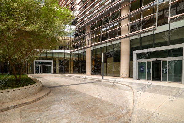 The image shows a modern building with a large glass facade and metal beams, featuring glass entrances. Trees and plants adorn the exterior courtyard, which includes tiled walkways. The overall environment conveys a sense of modernity and organization.