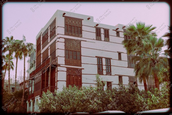 An image of a large, traditional building with four stories, featuring Islamic and Arab architectural style with prominent wooden windows and distinct paneling on the white walls. The building is surrounded by several palm trees, giving it a tropical appearance, and the façade is covered with dark brown wood. The sky in the background is clear with a light pink hue.