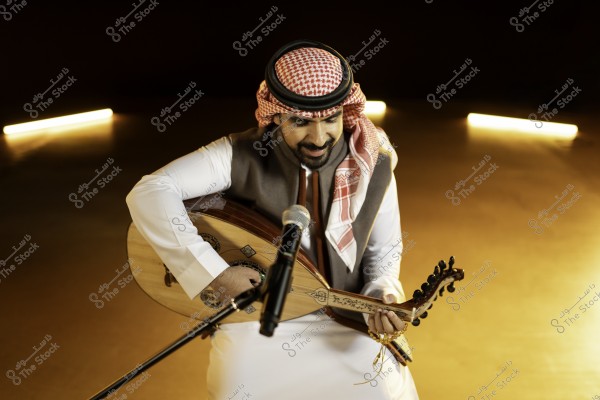 رجل يرتدي الزي التقليدي يعزف على آلة العود أمام ميكروفون في مكان مظلم بإضاءة دافئة.