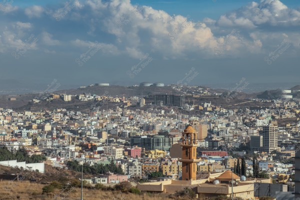 A panoramic view of a city sprawling over hills with multi-story buildings in various colors. In the foreground, there is a mosque\'s minaret in a light brown color. The background features hills with large storage tanks and additional buildings. The sky is cloudy.