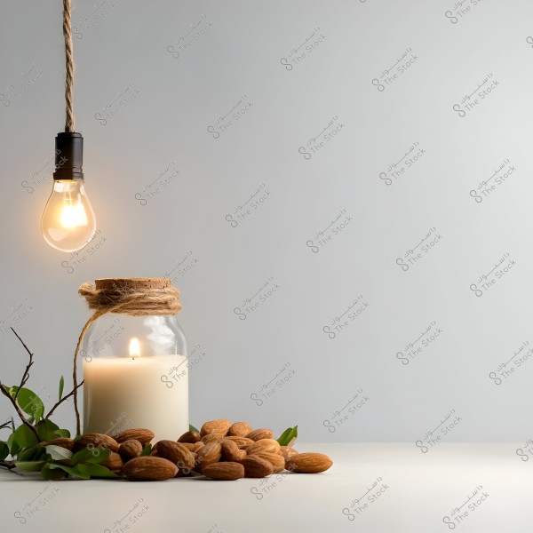 Image showing a hanging glowing bulb with a rope-wrapped wire on the left, and on the table there is a large white candle placed inside a glass jar, wrapped with rope at the top. The jar is surrounded by almonds and small green branches. The background is a simple gray.