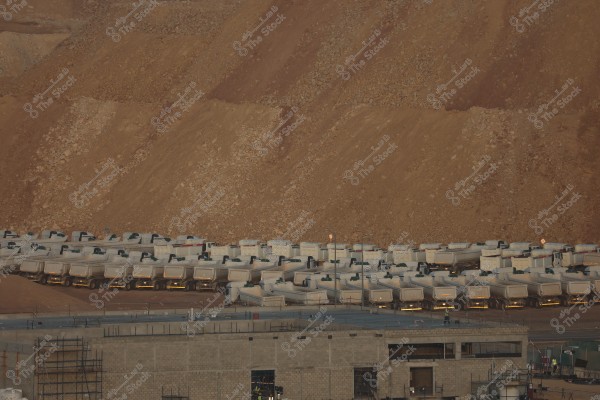 A group of large white trucks lined up in rows in front of massive dirt hills, in an area that might be designated for industrial or construction purposes. In the foreground, a building under construction is visible with scaffolding and ongoing construction work.