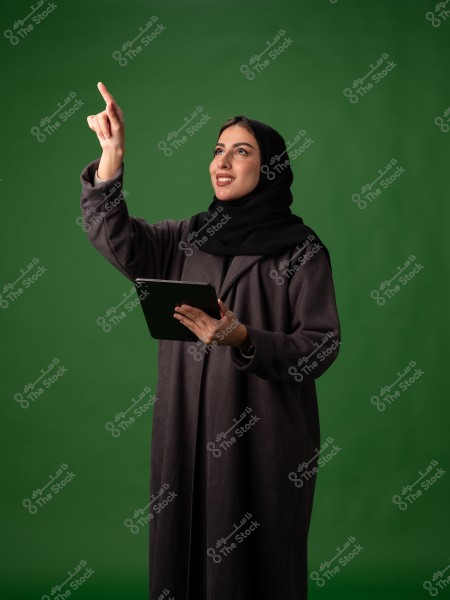A portrait of a woman wearing a black abaya and hijab, standing against a green background. She is holding a tablet in one hand and using the other to point upwards. Her expression appears welcoming and focused on something above.