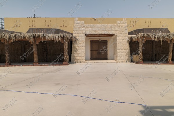 The image shows an outdoor courtyard with a building in a traditional style, featuring stone walls and decorative elements on the upper parts of the walls. In the center, there is a large wooden door with a traditional design, flanked by canopies made of palm fronds with wooden pillars. The front courtyard is wide and open.