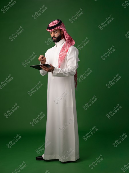 A man wearing traditional Saudi attire with a headscarf and agal, standing against a green background, looking at a tablet in his hand.