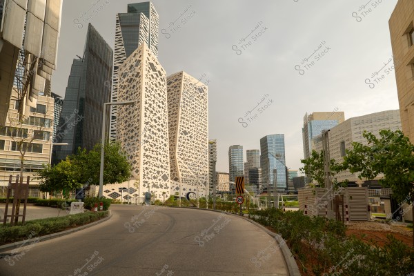 An image of a modern cityscape featuring a cluster of high-rise buildings with distinctive architectural designs, set in an area filled with trees and greenery. The central structure stands out with its unique geometric patterns and integrated windows. The sky is overcast, adding a dramatic effect to the scene.