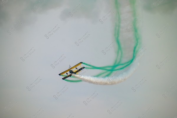 A biplane flying in the sky with green and white smoke trailing behind it, adding a dramatic effect to the aerial display. The background features the sky with some light gray clouds.