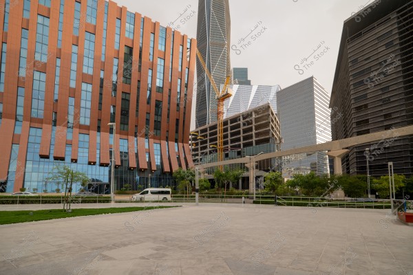 Image of an urban scene in a modern city. A modern building with a brown facade and a tall glass tower in the background are visible. There\'s a building under construction with a yellow crane in the center. An open space with a walkway surrounded by green trees and a parked white vehicle are also visible.