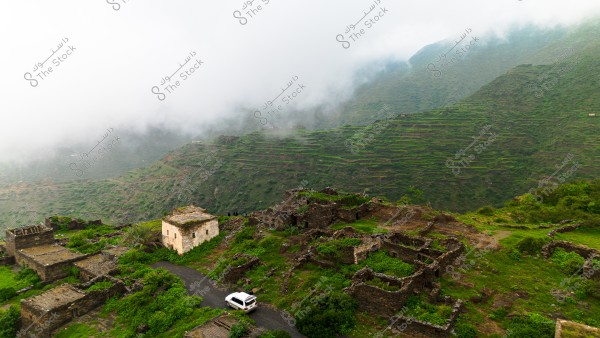 A scenic view of an old traditional village in a mist-covered green mountainous area. The image shows ancient brown buildings scattered on the lush slopes, with a small white vehicle parked on the path leading to the village. Low clouds touch the mountain peaks, adding an aura of mystery and charm to the scene.