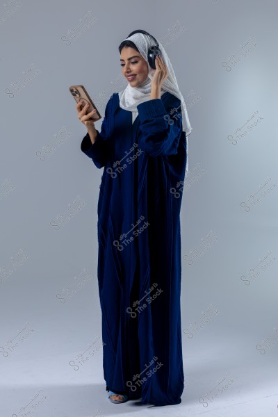 صورة لامرأة ترتدي عباءة زرقاء وحجاب أبيض تقف في استوديو. تمسك بيدها اليمنى هاتفًا محمولًا وبيدها اليسرى تضع سماعات الرأس على أذنيها. تبدو مبتسمة وتنظر إلى الهاتف. الخلفية باللون الرمادي الفاتح.