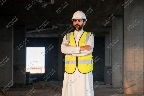 An image of a person standing inside a construction site wearing a traditional white thobe, a yellow safety vest, and a white hard hat. The person stands with arms crossed, displaying a serious expression. Part of the building\'s exterior is visible through an opening in the wall.