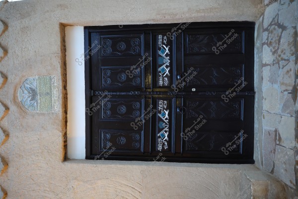 An image of a dark wooden door adorned with intricate carvings and traditional motifs, possibly featuring Arabic script in white at the center. The door appears to be part of a mud-brick wall with some stonework on the right. A carved stone plaque is mounted on the wall next to the door.