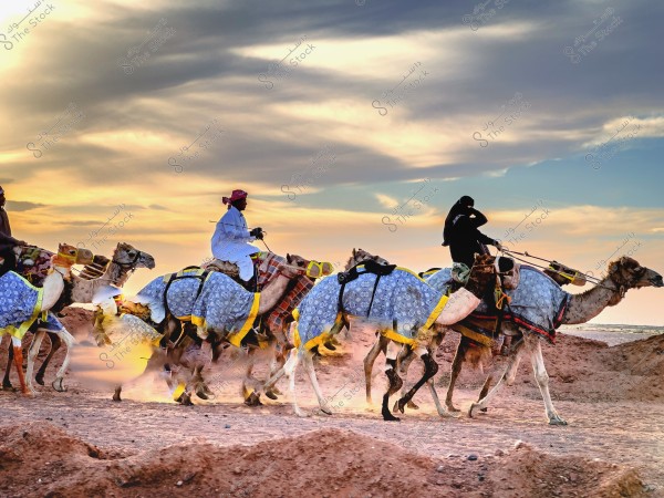 مجموعة من الأشخاص يرتدون ملابس تقليدية, يمتطون جمالًا مغطاة بأغطية مزخرفة, يتسابقون في الصحراء خلال غروب الشمس. السماء مزينة بالغيوم الملونة وخلفية من الرمال الصفراء.