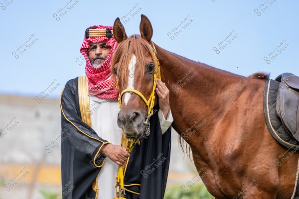 صورة لشخص يرتدي ثوبًا عربيًا تقليديًا مع الشماغ والغترة, يقف بجانب حصان بني ويربط بيده باللجام. الشخص يرتدي عقالًا أسود فوق الغترة البيضاء والشماغ الأحمر. الحصان مزود بسرج وجميع التفاصيل تبرز في بيئة خارجية تحت السماء الصافية.