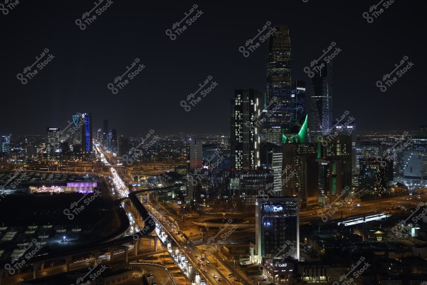 A nighttime view of Riyadh, Saudi Arabia, showcasing a cluster of illuminated skyscrapers, including the iconic Kingdom Tower. The city is vibrant with streetlights and busy highways filled with cars. The buildings feature modern and innovative architectural designs.