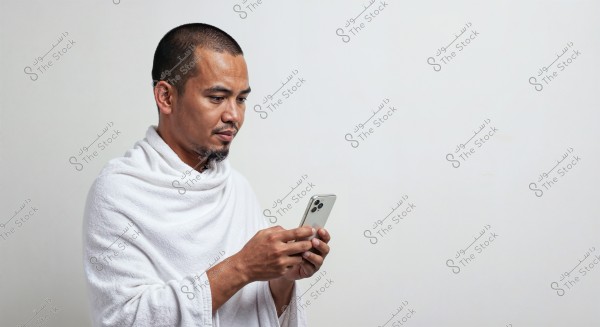 A portrait of a man looking at a smartphone in his hand. The man is wearing a white seamless garment, which suggests it may be related to traditional attire used for Hajj or Umrah in Arab countries. The background is plain white, emphasizing the focus on the man and his phone.
