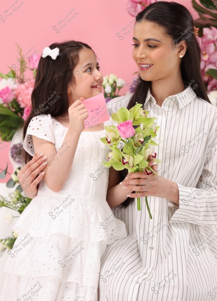 An image of a woman and a little girl seated against a pink background. The girl is wearing an elegant white dress with a white bow in her hair, holding a pink card with \"To Mom\" and a heart drawn on it. The woman is wearing a white and black striped shirt and holding a bouquet of flowers with a pink rose and green orchids. They both look at each other and smile warmly.
