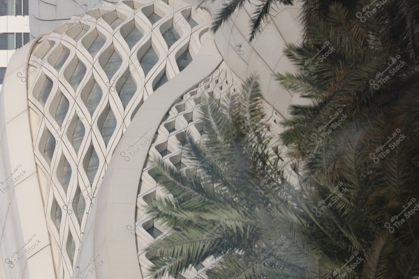 An image depicting a complex architectural façade with a wavy geometric design, overlaid at the front by green palm leaves. The building features interlocking triangular patterns that shine with soft lighting, suggesting a modern architectural style. In the background, there is another building with glass paneling visible.