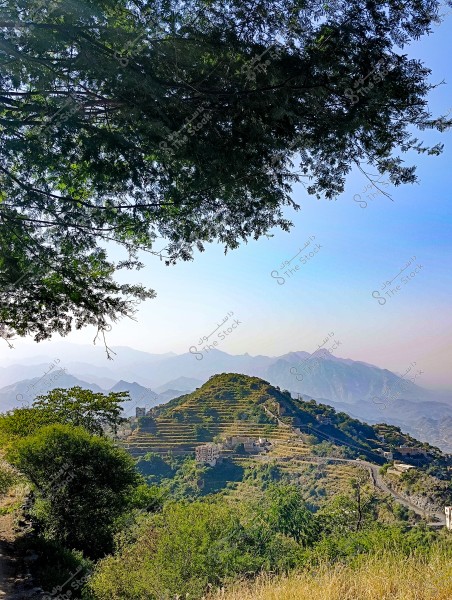 A mountain landscape of a village in Yemen, featuring green agricultural terraces on a mountain range. The village has traditional buildings scattered among the terraces. In the foreground, dense trees are visible, while in the background, more mountains can be seen under a clear blue sky.