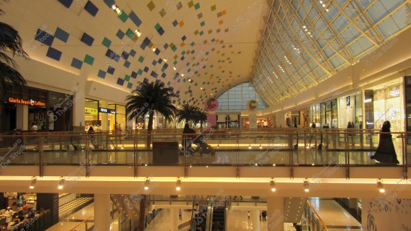 The image shows an interior view of a spacious shopping mall with two floors of retail stores. Palm trees are placed as part of the indoor decor. The ceiling features colorful geometric patterns, and large glass windows on the right allow natural light to flood in. People can be seen walking and shopping on the upper level, with signs for various shops visible.