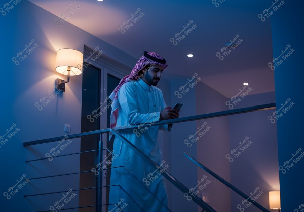 An image of a man standing by an interior staircase railing in a modern home, wearing a white thobe and a red-checkered headscarf, looking at a mobile phone. The lighting is dim with blue and white tones and a wall lamp illuminated in the background.