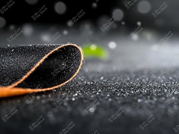 A close-up image of a flat surface covered with black sandpaper adorned with tiny particles resembling diamonds or sand. The paper is slightly curled, revealing its distinctive orange edge. The background is blurred and soft with scattered light bokeh.