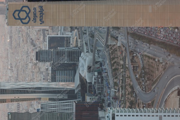 View of a bridge and pedestrian walkway in Riyadh, Saudi Arabia, surrounded by skyscrapers and highways. The image features a tall building with the "SABIC" logo, alongside traffic flow on the highway and a bridge adorned with modern and intricate architectural design in the city center.