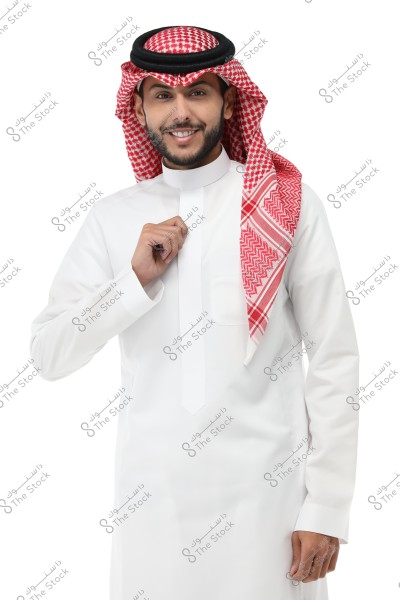 A portrait of a man wearing a traditional white thobe with a red and white checkered ghutra and agal. He is smiling and looking at the camera against a white background. The image shows him from the chest up.