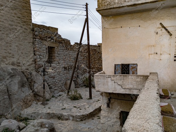 An image of an old village featuring stone buildings and weathered walls. The cream-colored house on the right has a small balcony with a blue window. The ground is paved with stones, and there are old-fashioned lamp posts. Large rocks are visible on the left. The sky is overcast, and the image includes power lines.