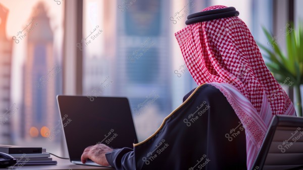 A man wearing traditional Gulf attire, including a keffiyeh and agal, is seated at his desk in front of a laptop. The desk is positioned near a large floor-to-ceiling window overlooking tall buildings. A green plant is visible in the right corner of the image.