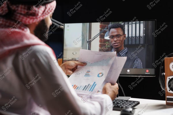 The image shows a man wearing traditional Gulf attire in white with a red checkered headscarf sitting in front of a computer, holding papers with charts. On the computer screen, another person appears in a video conference, wearing glasses and a striped shirt. Speakers and a keyboard are visible on the desk.