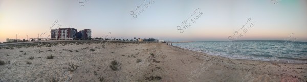 Panoramic view of a sandy beach at sunset, with the sea stretching calmly on the right. On the left side, a cluster of modern residential buildings is visible with dim lighting, surrounded by palm trees and street lamps. The sky displays warm sunset colors over the sea and buildings.