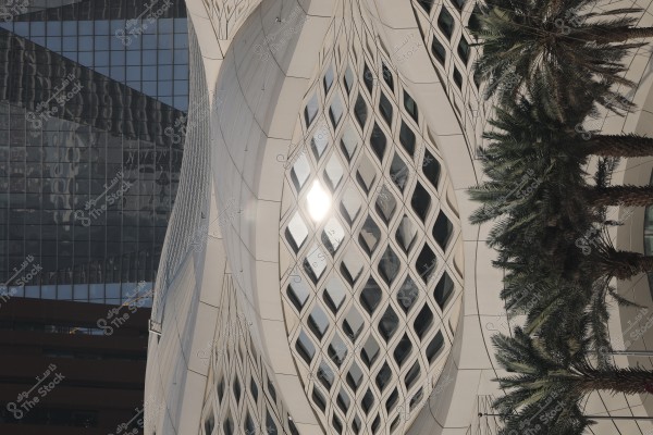 A modern building with a unique architectural design featuring glass facades and white geometric patterns. The image shows the sun reflecting on one of the glass surfaces, and palm trees are visible next to the building.