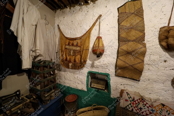 The image shows a traditional room with white and green painted walls. A decorative leather bag with geometric shapes and another smaller bag hang on the wall. A traditional white garment is hung on the left wall. At the bottom, there is a basket, some old tools, and a wooden crate. A small window in the wall contains an old radio. There is also a pillow decorated with traditional colors and patterns.