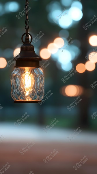 A glass lantern hanging from a metal chain, featuring faceted designs that emit a warm glow. The background shows circular bokeh lights in warm colors, with a blurred natural setting.