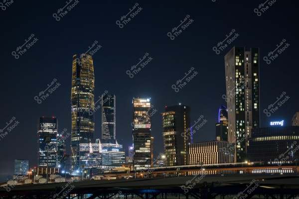 منظر لمجموعة من ناطحات السحاب في مدينة حديثة ليلاً. تظهر المباني مضاءة بأضواء ساطعة بألوان مختلفة, تبرز تفاصيل الهندسة المعمارية الحديثة. السماء داكنة توحي بجو هادئ, وبعض الأضواء في الأفق تظهر حركة الحياة في المدينة.