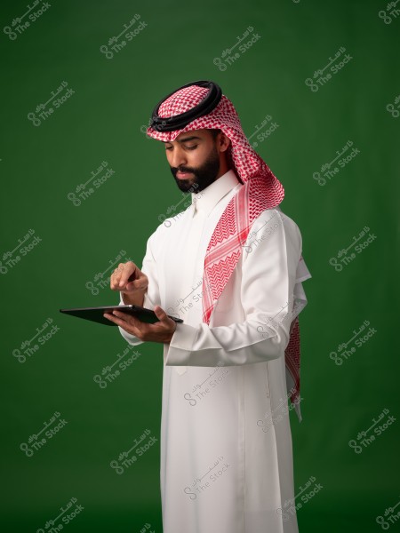 A portrait of a man wearing traditional Saudi attire and a keffiyeh stands in front of a green background using a tablet with his hands.